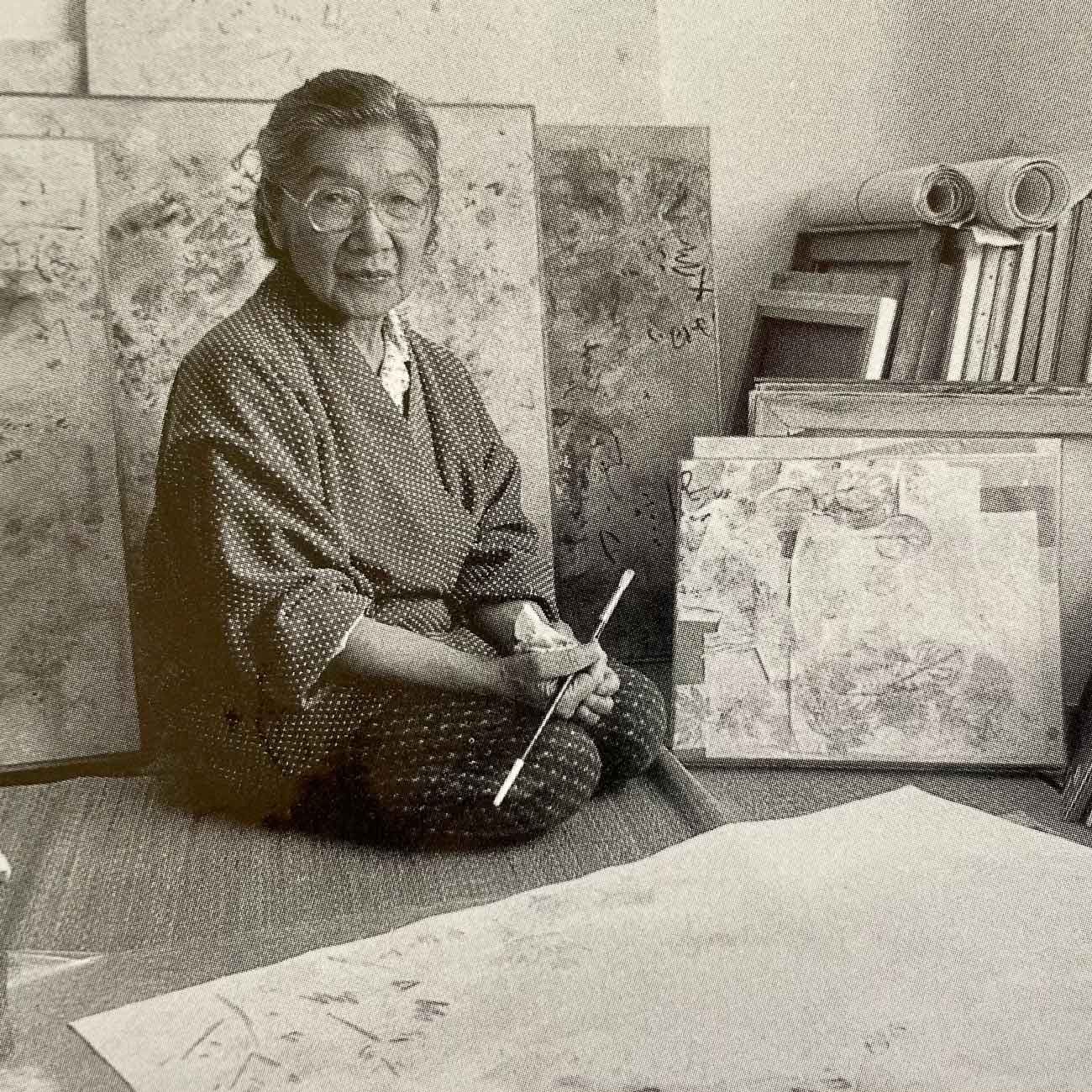 A black and white photograph of an elderly Hisako Hibi sitting on the floor of her workshop surrounded by her paintings, holding a paint brush while looking at the camera. 