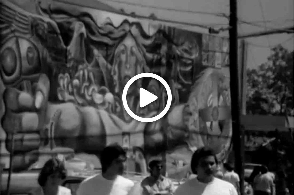 A video still showing people in front of a large mural. 