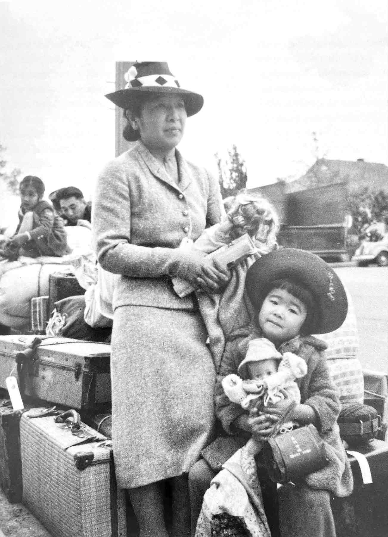 A black and white photo by Dorothea Lange title: “Hisako and Ibuki Hibi with doll awaiting the bus to Tanforan,”