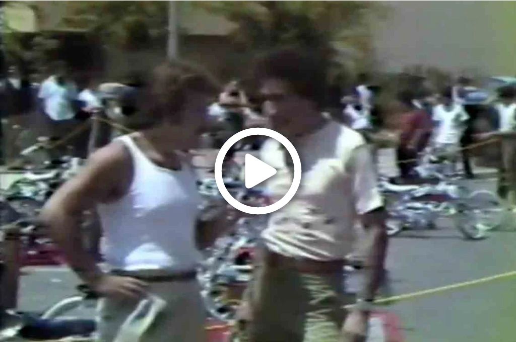 A video still of two men talking in the street, in front of parked motor cycles.