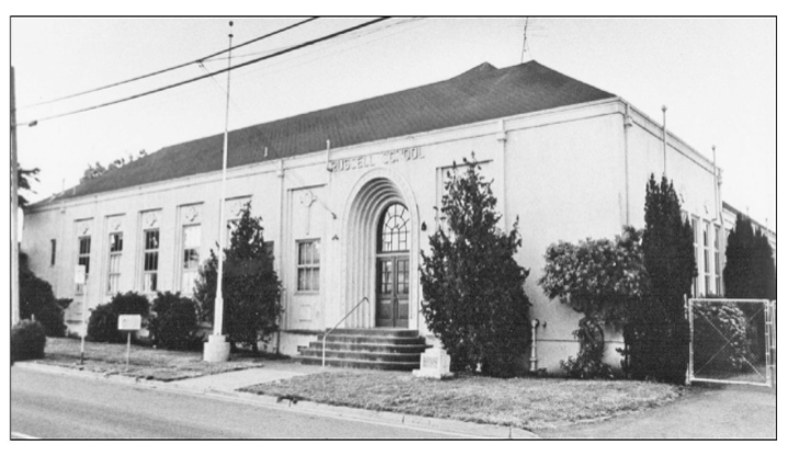 Black and white photo of a large white building with a tall door and tall windows. Above the door there is a sign saying "Russell School" in large letters.