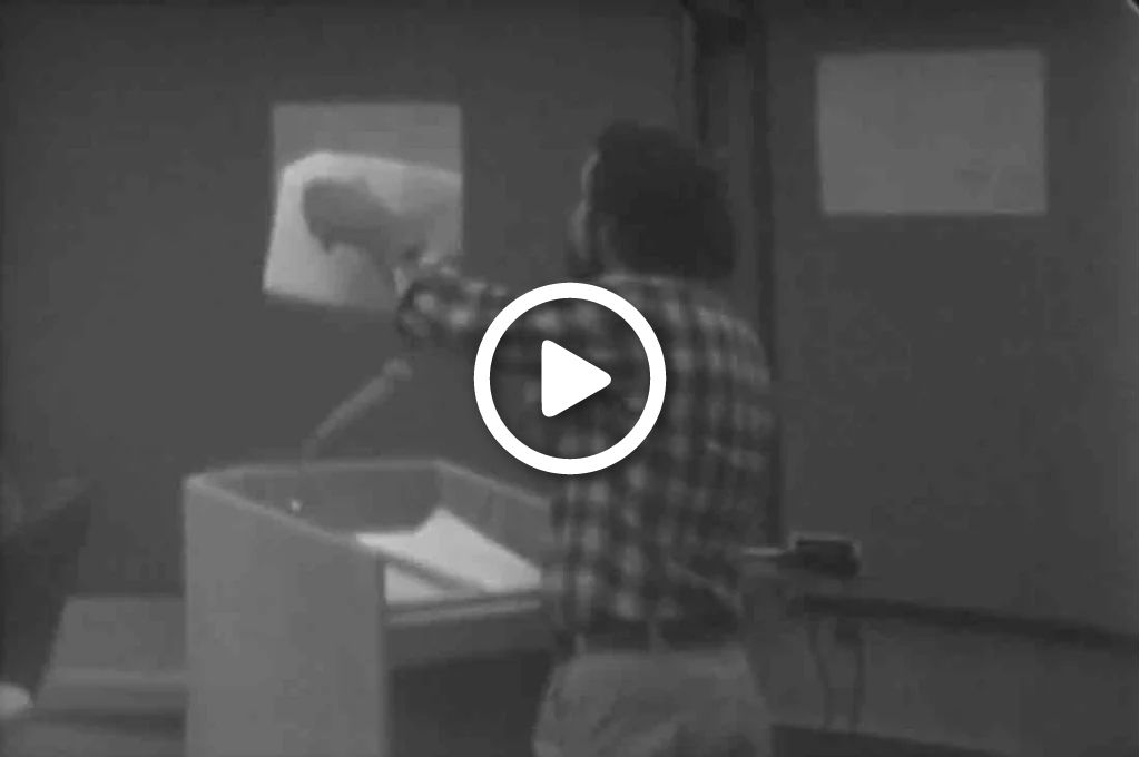 A Video still of a man standing holding up some documents at a lectern at a city council meeting. 